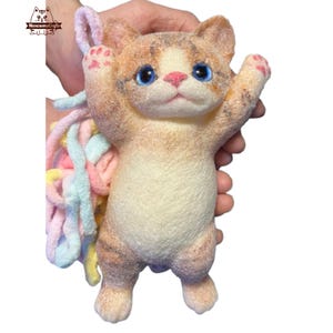 May include: A small, felted cat toy with a light brown and white fur pattern. The cat has blue eyes and a pink nose. The toy is being held by a hand, and a colorful yarn tail is attached to the cat's back.