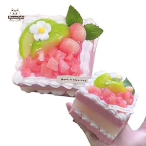 May include: Two decorative cake slices with pink and white frosting, topped with green and pink fruit, and a white flower. The cakes have the text "Have a nice day" on a small white label.