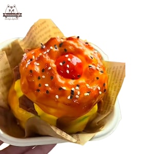 May include: A miniature burger with a glossy orange glaze, sesame seeds, and a red cherry on top. The burger sits in a white container lined with brown paper. The logo "MeowsomeCrafts" is in the upper left corner.