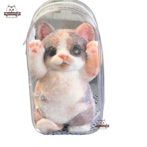 May include: A small, white, gray, and orange cat figurine is inside a clear plastic backpack. The cat is standing on its hind legs with its front paws raised. The backpack has a zipper on the top.