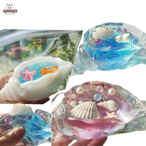 Può includere: Tre contenitori di plastica trasparente riempiti di slime colorato e varie decorazioni a tema marino. I contenitori hanno la forma di conchiglie e contengono oggetti come una stella marina, un granchio, una conchiglia e una nuvola bianca.