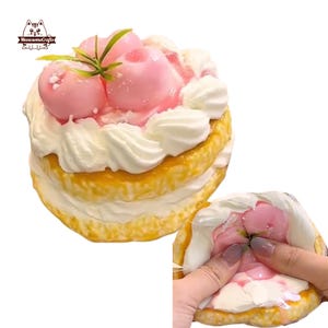 May include: A layered cake with white frosting and pink peach toppings. The cake has a yellow base and is topped with three pink peach-shaped decorations and green leaves. The cake is being squeezed, revealing the soft interior.