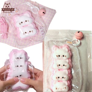 May include: A pink and white cat-shaped squishy toy with a bell charm. The toy is packaged in a clear plastic bag with a pink and white checkered ribbon.