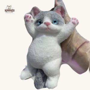 Puede incluir: Figura de gato de fieltro hecha a mano en una pose juguetona, con los brazos levantados. El gato tiene un cuerpo blanco, marcas grises, ojos azules y patas y nariz rosas. La figura es un artículo artesanal único.