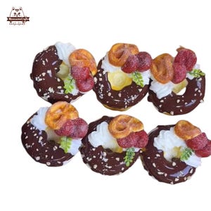 May include: Six chocolate-covered donuts with white frosting, pretzel-shaped toppings, and red fruit decorations. The donuts are arranged in two rows of three, showcasing a dessert theme.