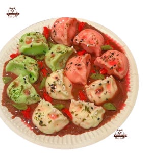 Può includere: Un piatto di ravioli colorati in salsa rossa. I ravioli sono verdi, rosa e bianchi, con spruzzi neri e bianchi. Il piatto è bianco con un bordo strutturato. L'immagine include il testo "Meowesome Crafts".