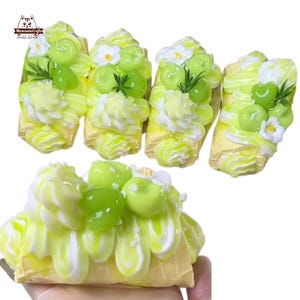 May include: A selection of miniature, decorative food items resembling grape-topped pastries. Each piece features a yellow base, topped with white and yellow frosting, green grapes, and small white flowers. The items are arranged on a white background.