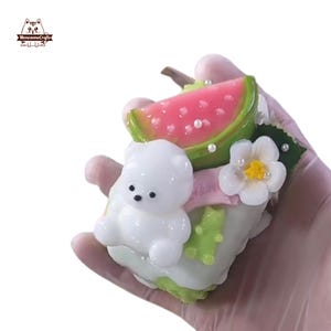 May include: A white bear figurine with a watermelon slice, a white flower, and green accents. The watermelon is pink with white seeds and a green rind. The flower has a yellow center. The figurine is held in a hand.