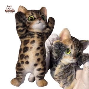 May include: Two cat figurines with green eyes and brown and black markings. One cat is standing with its paws raised, while the other is held by a gloved hand. The figurines have a detailed, realistic appearance, with a playful pose.