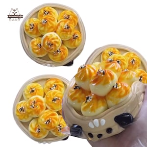 Può includere: Miniature di panini bao, con vortici marrone dorato e semi di sesamo nero, presentate in cestini vapore di bambù e un contenitore decorativo. Articoli artigianali a tema cibo.