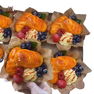 May include: A collection of miniature, decorative bread loaves, croissants, and pastries, artfully arranged with faux berries and cream. The baked goods are golden brown, and the berries are red and blue. The arrangement is presented on paper.