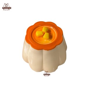 May include: A white and orange miniature dessert shaped like a pumpkin with three yellow corn kernels on top.
