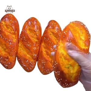 Può includere: Quattro pagnotte di pane a forma di pretzel color oro con semi di sesamo. Una pagnotta è tenuta in mano da un guanto. Il pane ha una finitura lucida e una superficie strutturata. Il logo "MeowesomeCrafts" è in alto a sinistra.