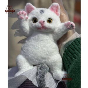 Puede incluir: Un peluche de gato blanco hecho a mano con orejas, patas y nariz rosas. El gato tiene ojos amarillos y una pequeña mancha negra en la cabeza. El peluche es suave y esponjoso, con los brazos extendidos.