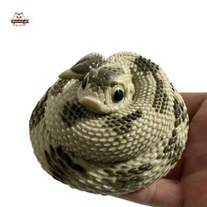 以下が含まれることがあります： リアルな手描きの巻き付けられたガラガラヘビの置物。ヘビは薄い茶色で濃い茶色の模様があり、大きく黒い目があります。