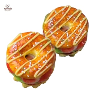 May include: Two decorative resin donuts with a glazed orange top, drizzled with white icing and sprinkled with sesame seeds. The donuts have a red and green filling, resembling a burger. The image is on a white background.