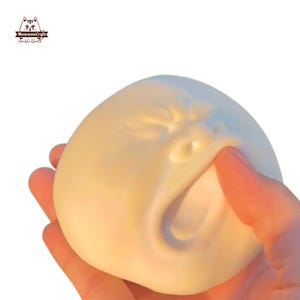 May include: A white squishy toy shaped like a face with a wide open mouth and a furrowed brow. The toy is being squeezed by a hand.