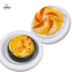 Può includere: Miniature di cibo su piatti bianchi. Un piatto contiene ravioli in salsa gialla, l'altro una piccola ciotola di zuppa con elementi arancioni e gialli. I cibi sono probabilmente per una casa delle bambole o un'esposizione in miniatura.