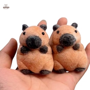 Può includere: Due giocattoli di peluche capibara marroni e neri con pelliccia soffice. I capibara sono seduti su una mano.