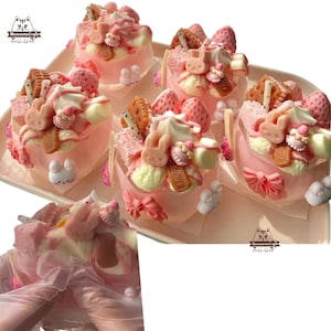 May include: Five pink and white mini cakes decorated with various toppings, including strawberries, chocolate, and sprinkles. The cakes are arranged on a white tray with a pink ribbon.