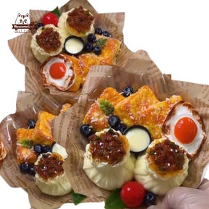 Pode incluir: Uma exibição de alimentos em miniatura, incluindo ovos fritos, bolinhos e rabanadas, dispostos em papel. Guarnecido com mirtilos e uma cereja vermelha. A comida é apresentada em papel castanho com texto impresso.
