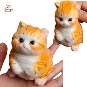 May include: Two small orange and white cat figurines. The cat on the left is sitting with its paws tucked under its body. The cat on the right is standing with its paws outstretched. Both cats have black eyes and pink noses.
