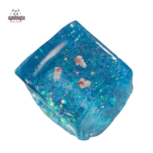 May include: A square, transparent resin paperweight with a blue, glittery interior. The paperweight contains three small, white and orange goldfish. The MeowesomeCrafts logo is in the upper left corner.