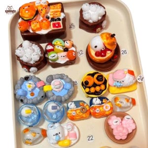 May include: A collection of miniature food-themed figurines, including burgers, sushi, and desserts, arranged on a white tray. The figurines feature bright colors and detailed designs, with some resembling cartoon characters. The tray is numbered with small black numbers.