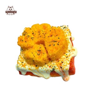 May include: A miniature food item resembling a piece of toast topped with a fried, breaded food item and a creamy white sauce. The toast is red, and the breaded food item is a golden yellow colour. The item is decorated with green flecks.