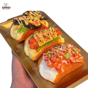 Può includere: Tre hot dog decorativi su un vassoio dorato. Gli hot dog hanno vari condimenti, tra cui quello che sembra essere relish, senape e altri ingredienti colorati. I panini sono di colore giallo chiaro e il vassoio è rettangolare.