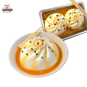 Può includere: Un panino bianco al vapore con semi di sesamo neri e un vortice di salsa arancione in una ciotola bianca con bacchette. Il panino è in una ciotola bianca con un vortice di salsa arancione. Ci sono altri due panini in un contenitore argentato con bacchette.