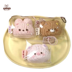 May include: Three crocheted animal-shaped coin purses: a pink bunny, a brown bear, and a pink pig. Each is packaged in a clear plastic pouch. The purses are displayed on a yellow, scalloped tray. The brand logo is in the top left corner.