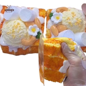 May include: A close-up of a dessert resembling a slice of cake, topped with a scoop of ice cream, marshmallows, nuts, and a white flower. The cake has a yellow and orange colour. The dessert is held in a clear plastic wrap.