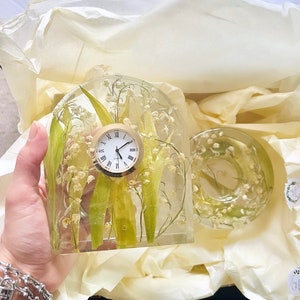 May include: A clear resin clock with a round face and Roman numerals, featuring lily of the valley flowers and green leaves. A matching round resin piece is also visible. The items are presented in a gift box with tissue paper.