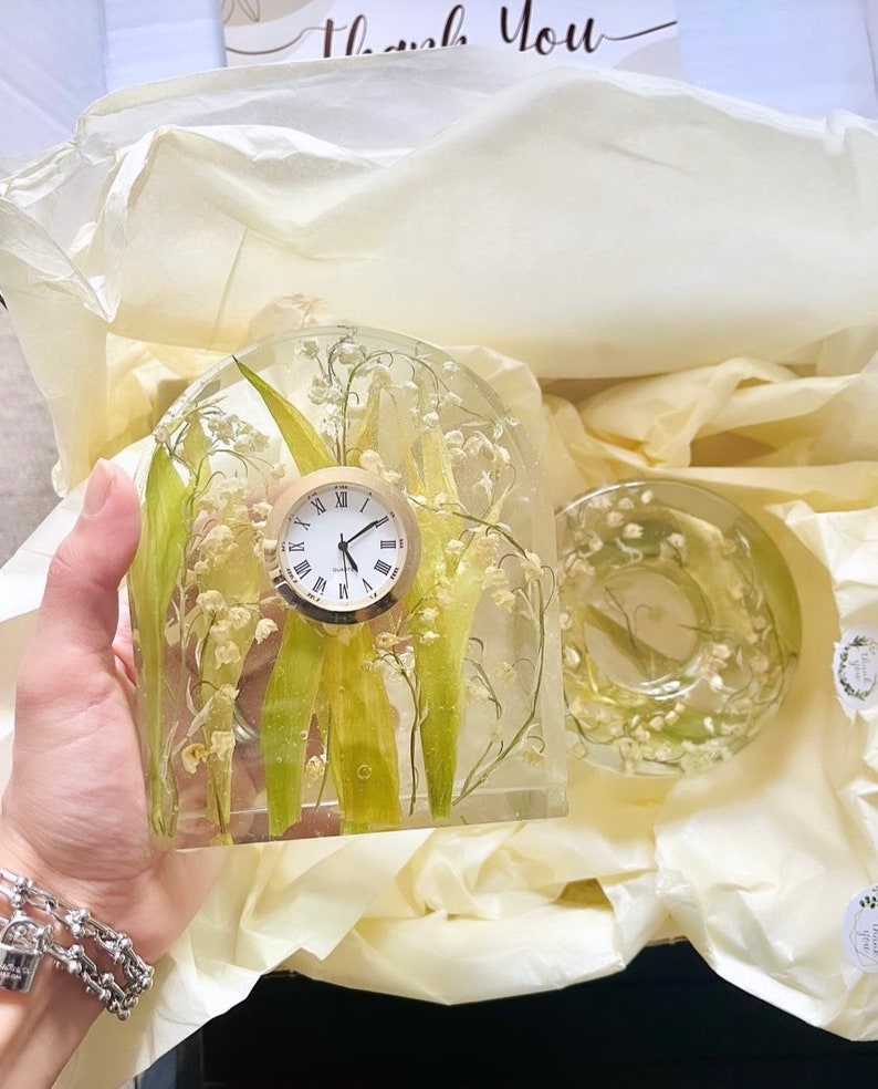 May include: A clear resin clock with a round face and Roman numerals, featuring lily of the valley flowers and green leaves. A matching round resin piece is also visible. The items are presented in a gift box with tissue paper.