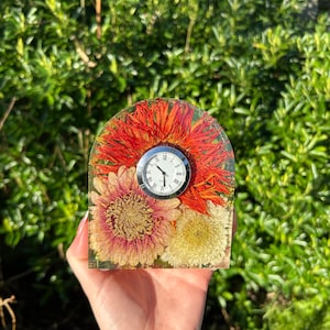 May include: A small, square clock encased in clear resin, featuring dried orange and pink flowers. The clock face is white with black Roman numerals and hands. The resin block is held in a hand against a backdrop of green foliage.