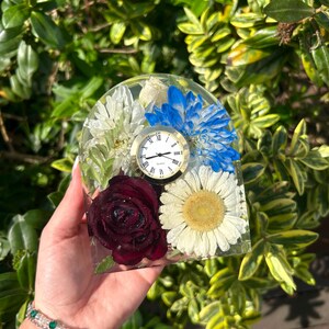 May include: A clear resin clock with a gold-tone clock face, Roman numerals, and a variety of preserved flowers. The flowers include a deep red rose, white daisies, and blue and white chrysanthemums. The clock is held in front of a green and yellow leafy background.