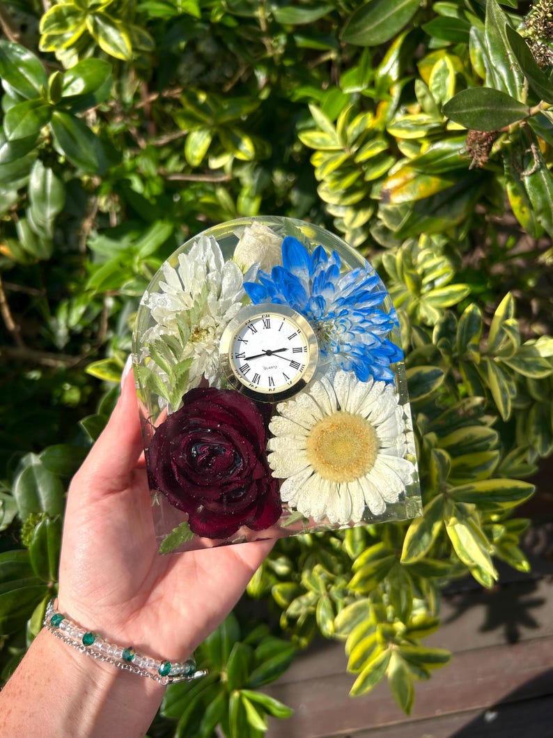 May include: A clear resin clock with a gold-tone clock face, Roman numerals, and a variety of preserved flowers. The flowers include a deep red rose, white daisies, and blue and white chrysanthemums. The clock is held in front of a green and yellow leafy background.