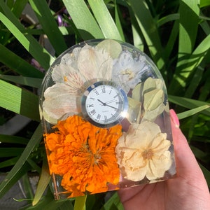 May include: A clear resin clock with a white clock face and Roman numerals. The clock is embedded with dried flowers in shades of white, orange, and cream. The clock is held in front of green foliage.