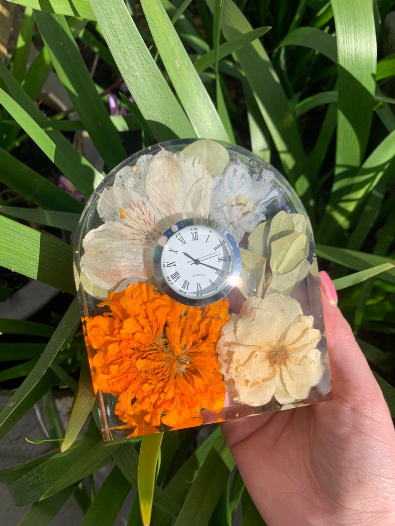 May include: A clear resin clock with a white clock face and Roman numerals. The clock is embedded with dried flowers in shades of white, orange, and cream. The clock is held in front of green foliage.