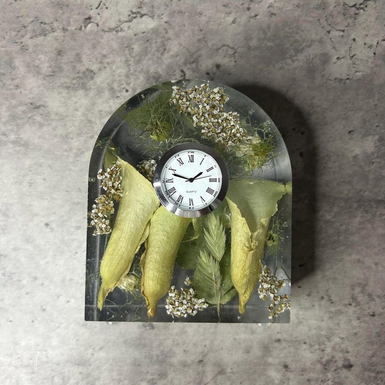 May include: A clear resin clock with a white clock face and black Roman numerals. The clock is embedded with dried leaves, flowers, and greenery. The clock is approximately 4 inches tall and 3 inches wide.