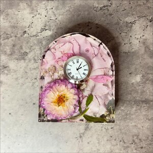 May include: A decorative clock with a silver frame and a white face with black Roman numerals. The clock is set in a clear resin base filled with pink flower petals and a purple and yellow flower.