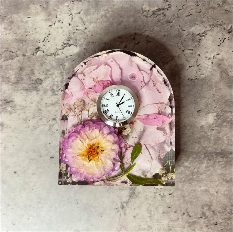May include: A decorative clock with a silver frame and a white face with black Roman numerals. The clock is set in a clear resin base filled with pink flower petals and a purple and yellow flower.
