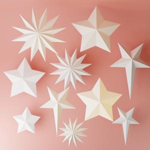 May include: An assortment of white paper star decorations in various sizes and designs, including multi-pointed and five-pointed stars. The stars are arranged on a soft pink background, creating a festive and decorative display.