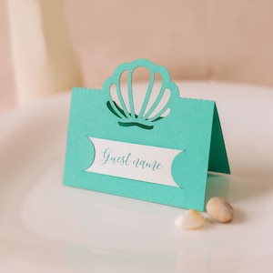 Può includere: Segnaposto blu turchese con un design a conchiglia ritagliato. La carta ha un'etichetta bianca con il testo "Guest name" stampato su di essa.