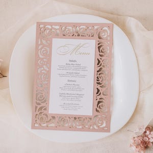 May include: A blush pink menu card with a laser-cut floral design, placed on a white plate. The menu features gold script text and a list of salads and entrees. The card is surrounded by a soft, neutral background.
