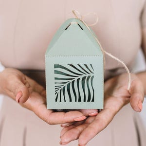 May include: A light green gift box with a cut-out design of a palm leaf. The box has a string attached to the top for tying.