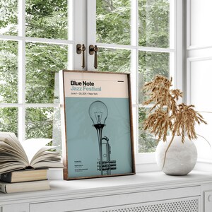 May include: A framed poster with a blue and white color scheme. The poster features a trumpet with a lightbulb on top and the text "Blue Note Jazz Festival" with the dates "June 1-30, 2021" and the location "New York".