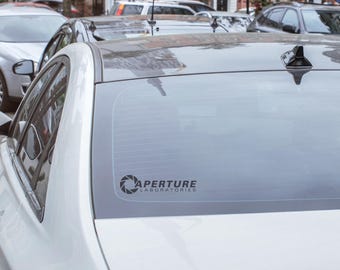 May include: A white car featuring a black Aperture Laboratories decal applied to the rear window. The decal displays a circular camera aperture icon next to the text Aperture Laboratories in a bold, sans-serif font, positioned on the glass.