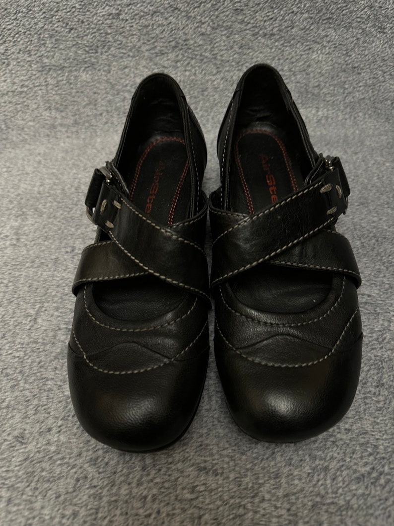 Może przedstawiać: Para czarnych sk&oacute;rzanych but&oacute;w z klamrą. Buty mają zaokrąglony nosek i niski obcas. Marka "Air Step" jest widoczna wewnątrz każdego buta.